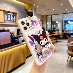May include: White phone case with a cartoon character design. The case features a black and white character with a pink face and a 3D figure on top. Purple and pink accents are visible.