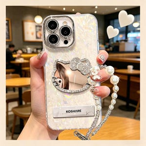 May include: A pearlescent white phone case with a Hello Kitty design, embellished with rhinestones and a pearl wrist strap. The case has a silver camera frame and the text "KOSHARE".