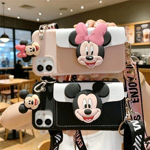 May include: Two phone cases featuring Mickey and Minnie Mouse. One case is pink with a Minnie Mouse design and a pink bow. The other case is black with a Mickey Mouse design. Both cases have a white flap and a strap.