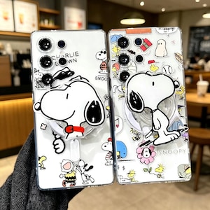 May include: Two clear phone cases featuring Snoopy and Charlie Brown designs. One case has Snoopy with a red collar and the text "CHARLIE BROWN". The other case has Snoopy with a flower and the text "SNOOPY". Both cases have a pop-up grip.