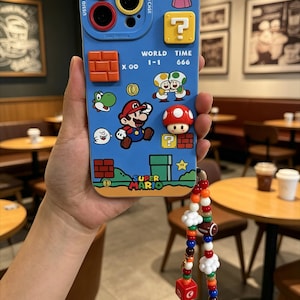 May include: A blue phone case featuring Super Mario characters and elements. The case includes a 3D brick block, a question mark block, and various character stickers. The case has a wrist strap with colorful beads and charms. The text "SUPER MARIO" is visible.