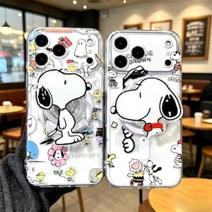 Puede incluir: Dos fundas transparentes para teléfono con Snoopy de Peanuts. Las fundas tienen un diseño de Snoopy blanco con un agarre emergente. Las fundas están decoradas con imágenes de dibujos animados de Snoopy, Charlie Brown y otros personajes.