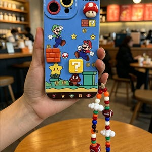 Puede incluir: Funda de teléfono azul con personajes de Super Mario, incluyendo a Mario, Luigi, un hongo y bloques de interrogación. La funda tiene un diseño 3D y un charm de cuentas de colores con una pequeña figura de Mario.