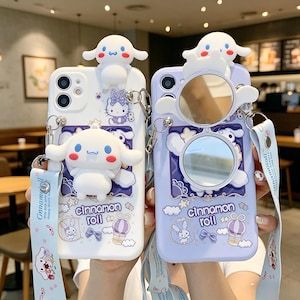 May include: Two phone cases, one white and one lilac, with a cartoon character design. The white case has a 3D character on top and a matching design. The lilac case has a mirror and a similar design. Both cases have a strap with text.