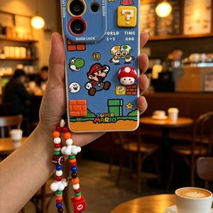 May include: A blue smartphone case featuring Super Mario characters, icons, and text including "GOOD LUCK", "WORLD 1-1", "TIME 666", and "SUPER MARIO". The case includes a colorful beaded phone charm with small cloud, cube, and character charms attached.