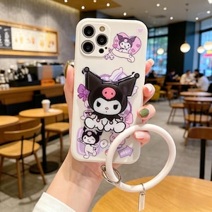 May include: White phone case featuring a cartoon character with a black head, pink accents, and a bow. The case has a matching white wrist strap with a silver-tone metal ring and a small tag that says "OPEN".