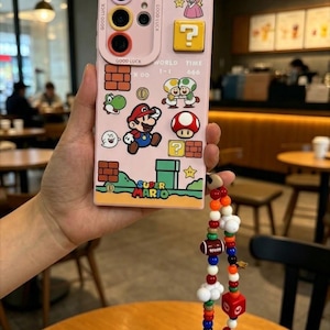 May include: Pink phone case decorated with Super Mario characters and game elements. The case has a decorative strap with colourful beads. The case includes the text "GOOD LUCK" and "SUPER MARIO".