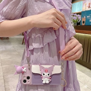 May include: A lavender phone case with a crossbody strap. The case features a cartoon character design with a white, purple, and pink color scheme. The case has a small pocket and a decorative charm.