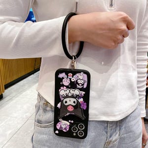 May include: Black phone case with a cartoon character design, featuring a black wrist strap. The case has a 3D design with a pig-like character, flowers, and other cartoon elements. The case has a camera cutout and a wrist strap.