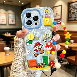May include: Clear phone case with Super Mario characters. The case has a blue border and features Mario, Princess Peach, coins, and question mark blocks. A beaded wrist strap with colourful beads and a yellow star charm is attached.