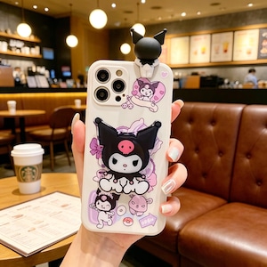 May include: A white phone case featuring a black and white cartoon character with a pink skull on its face, and a 3D figure on top. The case is decorated with other cartoon characters, flowers, and hearts. The word "OPEN" is visible.