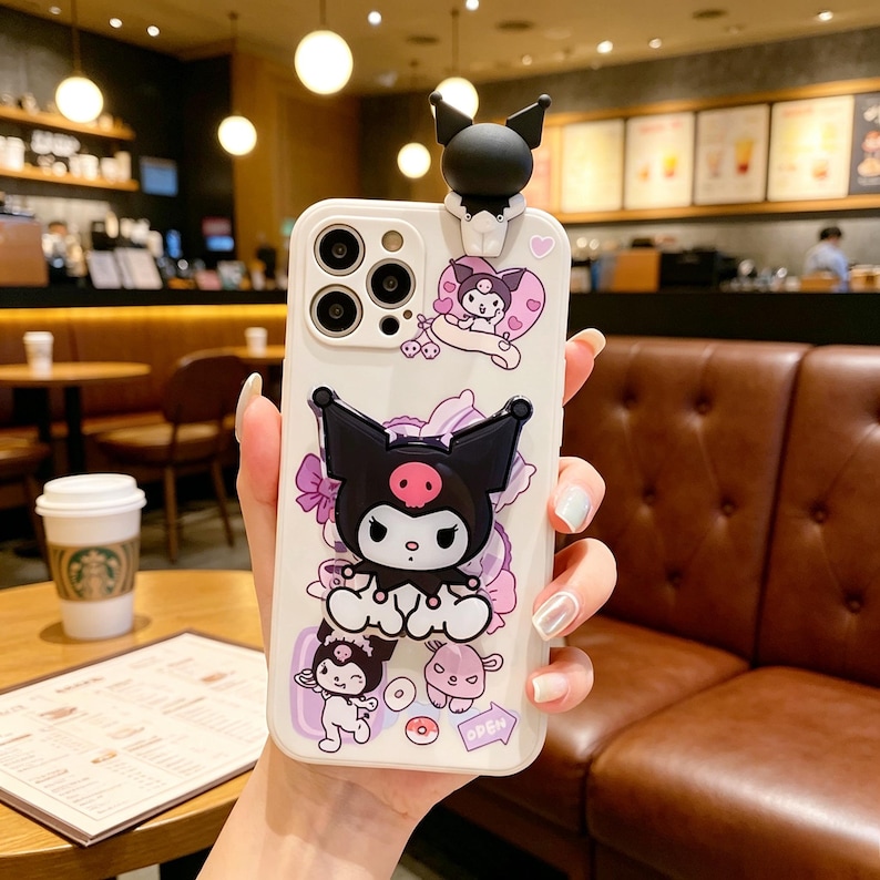May include: A white phone case featuring a black and white cartoon character with a pink skull on its face, and a 3D figure on top. The case is decorated with other cartoon characters, flowers, and hearts. The word "OPEN" is visible.