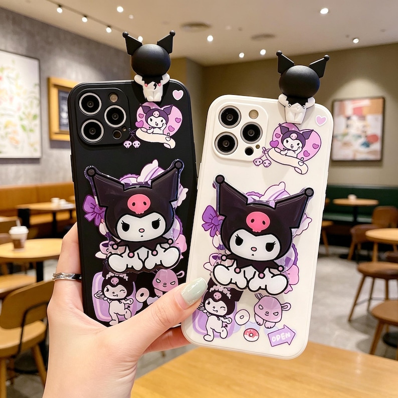 May include: Black and white phone cases featuring a cartoon character with large ears, a pink skull, and a bow. The cases have a 3D black ear design at the top. The character is surrounded by purple bows and other cartoon elements.
