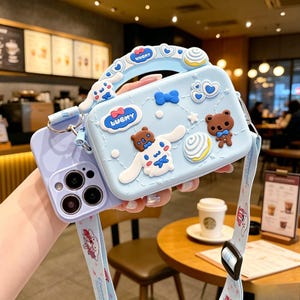 Peut inclure: Un étui de téléphone bleu clair avec un sac à bandoulière assorti. Le sac présente des motifs d'ours et de chiens de dessin animé, avec le mot "LUCKY" sur un nœud. La sangle et la poignée sont bleu clair. L'étui du téléphone est violet.