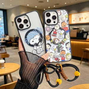 Puede incluir: Dos fundas transparentes para teléfono con bordes negros. Una presenta a Snoopy con la palabra "DING!!" en un bocadillo. La otra funda muestra varios personajes de Peanuts. Ambas fundas tienen un cordón negro y marrón.