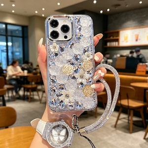 Könnte beinhalten: Transparente Handyhülle mit floralem Design, mit weißen und goldenen Rosen, klaren Kristallen und einer Strass-Handschlaufe. Die Hülle wird vor einem verschwommenen Café-Hintergrund gehalten.