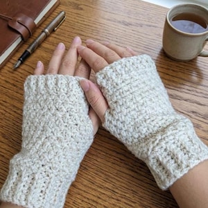 May include: A pair of cream-colored fingerless gloves featuring a textured crochet stitch pattern. The gloves have a ribbed cuff at the wrist and an open thumb hole, covering the hand from the knuckles down to the base of the wrist.