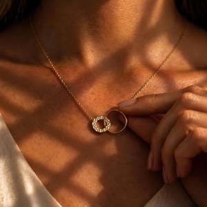 Puede incluir: Un collar de oro con dos círculos entrelazados. Un círculo está adornado con pequeñas piedras brillantes, el otro es una banda de oro lisa. El collar se muestra en el cuello de una persona, con su mano sosteniendo el colgante.
