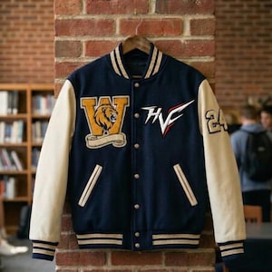 Puede incluir: Chaqueta universitaria azul marino con mangas color crema. Presenta un emblema de león, las letras "W" y "HVF", y el número "24". La chaqueta tiene cierre de botones y detalles a rayas en el cuello, puños y dobladillo.