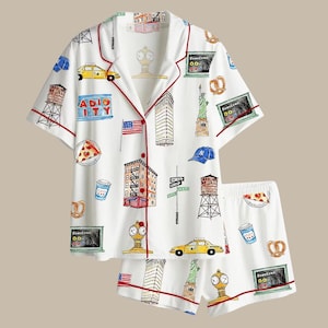 Pode incluir: Conjunto de pijama branco com tema de Nova York. A camisa de manga curta e os shorts apresentam um padrão repetido de marcos icônicos como a Estátua da Liberdade, táxis amarelos e o Empire State Building. O conjunto é adornado com debrum vermelho.