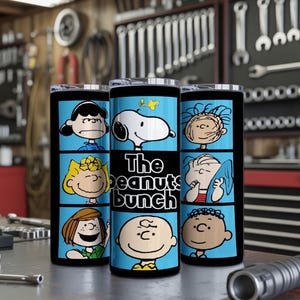 Puede incluir: Un vaso cil&iacute;ndrico con un estampado gr&aacute;fico azul y negro de personajes de Peanuts, incluyendo a Snoopy, Charlie Brown, Lucy y Linus. El texto "The peanuts bunch" est&aacute; impreso en blanco en el centro del vaso. Tiene una tapa transparente.