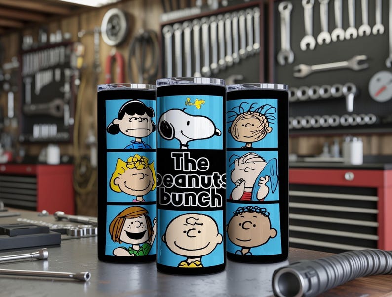 Puede incluir: Un vaso cil&iacute;ndrico con un estampado gr&aacute;fico azul y negro de personajes de Peanuts, incluyendo a Snoopy, Charlie Brown, Lucy y Linus. El texto "The peanuts bunch" est&aacute; impreso en blanco en el centro del vaso. Tiene una tapa transparente.