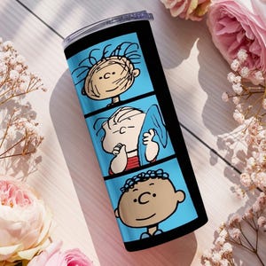 Puede incluir: Un vaso cil&iacute;ndrico de acero inoxidable con un dise&ntilde;o de tira c&oacute;mica vertical sobre un fondo azul brillante. El gr&aacute;fico muestra tres paneles distintos, cada uno de los cuales presenta a un personaje diferente de la serie de c&oacute;mics Peanuts.