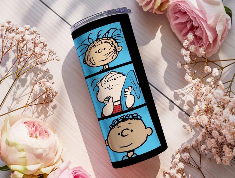 Puede incluir: Un vaso cil&iacute;ndrico de acero inoxidable con un dise&ntilde;o de tira c&oacute;mica vertical sobre un fondo azul brillante. El gr&aacute;fico muestra tres paneles distintos, cada uno de los cuales presenta a un personaje diferente de la serie de c&oacute;mics Peanuts.