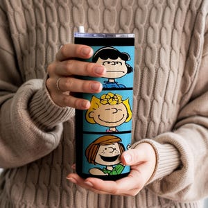 Puede incluir: Un vaso cil&iacute;ndrico de acero inoxidable con un fondo azul brillante y tres paneles horizontales apilados. Cada panel muestra una colorida ilustraci&oacute;n de un personaje de la tira c&oacute;mica Peanuts, incluyendo a Lucy, Sally y Peppermint Patty.