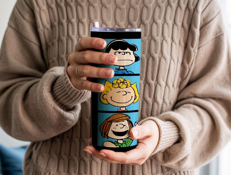 Puede incluir: Un vaso cil&iacute;ndrico de acero inoxidable con un fondo azul brillante y tres paneles horizontales apilados. Cada panel muestra una colorida ilustraci&oacute;n de un personaje de la tira c&oacute;mica Peanuts, incluyendo a Lucy, Sally y Peppermint Patty.