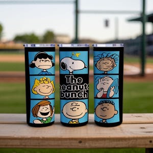 Puede incluir: Tres vasos cil&iacute;ndricos con tapas transparentes, con fondo azul claro y bordes negros. Los vasos muestran varios personajes de Peanuts en paneles estilo c&oacute;mic, con el texto "The peanuts bunch" impreso en el vaso central.