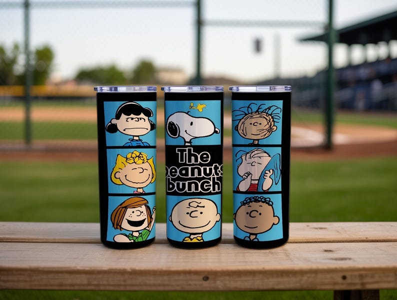Puede incluir: Tres vasos cil&iacute;ndricos con tapas transparentes, con fondo azul claro y bordes negros. Los vasos muestran varios personajes de Peanuts en paneles estilo c&oacute;mic, con el texto "The peanuts bunch" impreso en el vaso central.