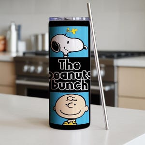 Puede incluir: Un vaso cil&iacute;ndrico de acero inoxidable con tapa transparente y pajita de metal. El exterior presenta un estampado gr&aacute;fico azul claro de Snoopy, Woodstock y Charlie Brown con el texto The peanuts bunch en letras blancas sobre fondo negro.