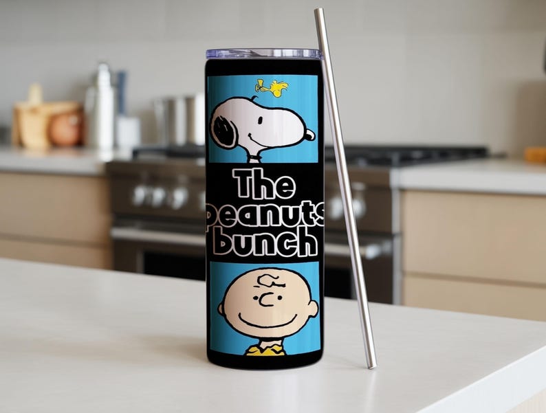 Puede incluir: Un vaso cil&iacute;ndrico de acero inoxidable con tapa transparente y pajita de metal. El exterior presenta un estampado gr&aacute;fico azul claro de Snoopy, Woodstock y Charlie Brown con el texto The peanuts bunch en letras blancas sobre fondo negro.