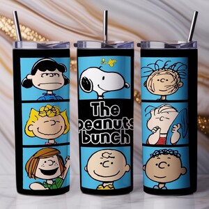 Puede incluir: Un vaso cil&iacute;ndrico de acero inoxidable con tapa transparente y pajita de metal, que presenta un estampado gr&aacute;fico azul de personajes de Peanuts como Snoopy y Charlie Brown, con el texto The peanuts bunch impreso en el panel central.