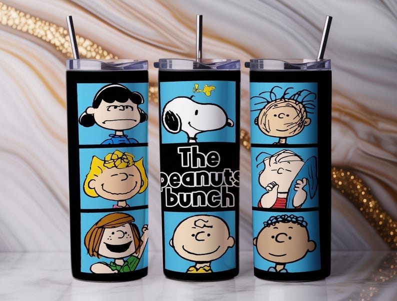 Puede incluir: Un vaso cil&iacute;ndrico de acero inoxidable con tapa transparente y pajita de metal, que presenta un estampado gr&aacute;fico azul de personajes de Peanuts como Snoopy y Charlie Brown, con el texto The peanuts bunch impreso en el panel central.