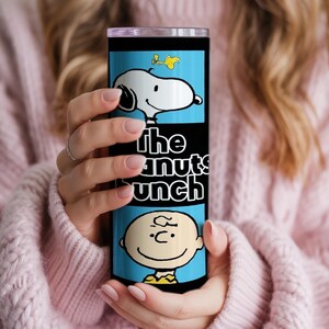 Puede incluir: Un vaso cil&iacute;ndrico de acero inoxidable con fondo azul claro y un estampado gr&aacute;fico de Snoopy, Woodstock y Charlie Brown. El texto "the peanuts bunch" est&aacute; impreso en letras blancas en el centro del vaso.