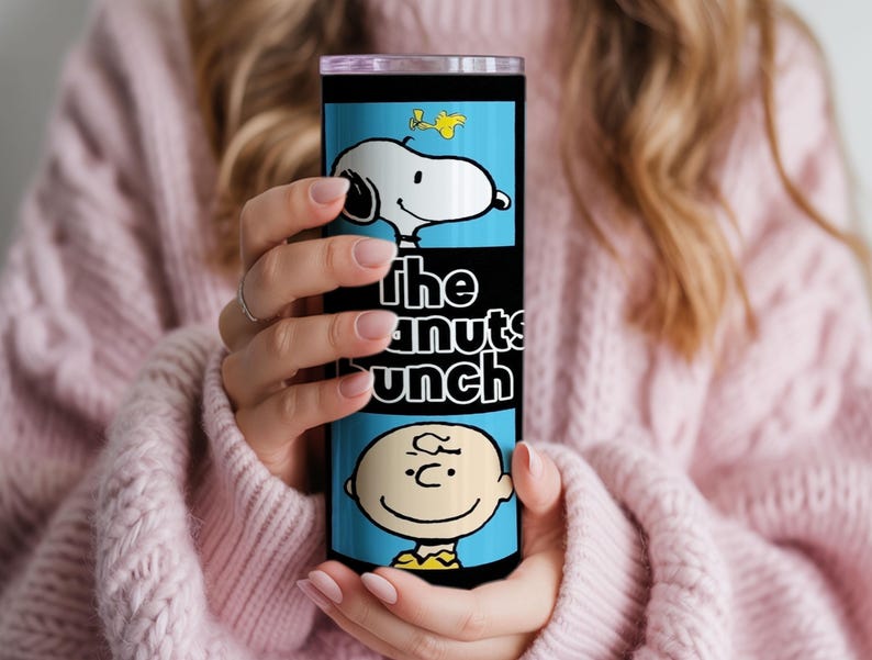 Puede incluir: Un vaso cil&iacute;ndrico de acero inoxidable con fondo azul claro y un estampado gr&aacute;fico de Snoopy, Woodstock y Charlie Brown. El texto "the peanuts bunch" est&aacute; impreso en letras blancas en el centro del vaso.