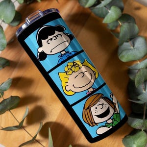 Puede incluir: Un vaso cil&iacute;ndrico de acero inoxidable con tapa de pl&aacute;stico transparente, que presenta un fondo azul brillante dividido en tres paneles horizontales. Cada panel muestra un personaje de la tira c&oacute;mica Peanuts, incluyendo a Lucy, Sally y Peppermint Patty.