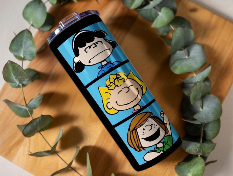 Puede incluir: Un vaso cil&iacute;ndrico de acero inoxidable con tapa de pl&aacute;stico transparente, que presenta un fondo azul brillante dividido en tres paneles horizontales. Cada panel muestra un personaje de la tira c&oacute;mica Peanuts, incluyendo a Lucy, Sally y Peppermint Patty.