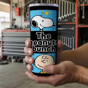 Puede incluir: Un vaso cil&iacute;ndrico de acero inoxidable con un dise&ntilde;o gr&aacute;fico en azul claro y negro. La secci&oacute;n superior muestra a Snoopy y Woodstock, el medio muestra el texto The Peanuts bunch en blanco y la parte inferior presenta a Charlie Brown.