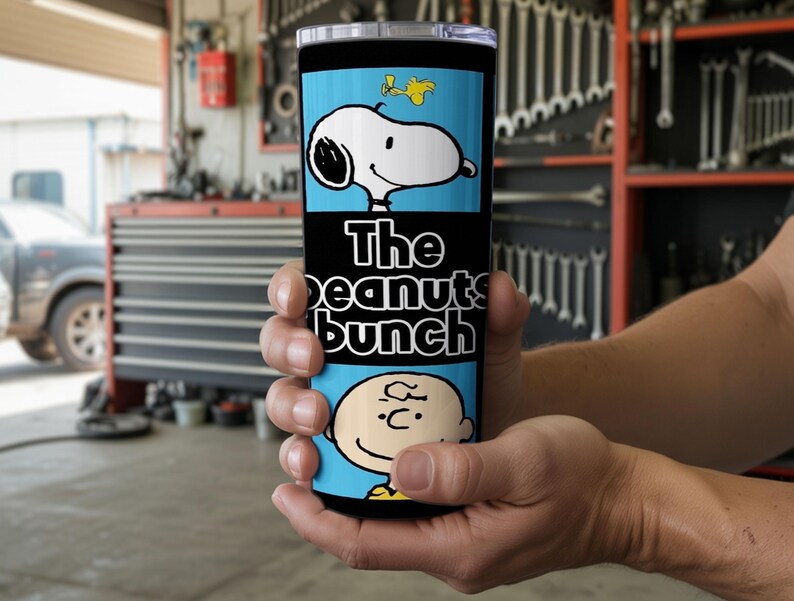 Puede incluir: Un vaso cil&iacute;ndrico de acero inoxidable con un dise&ntilde;o gr&aacute;fico en azul claro y negro. La secci&oacute;n superior muestra a Snoopy y Woodstock, el medio muestra el texto The Peanuts bunch en blanco y la parte inferior presenta a Charlie Brown.