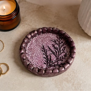 May include: A round, shallow trinket dish with a metallic purple, scalloped edge. The interior features a light purple base with a dark purple stippled texture and a prominent, dark purple embossed fern leaf design across the surface.