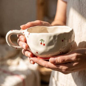 Könnte beinhalten: Cremefarbene Keramiktasse mit rotem Kirschmuster. Die Tasse hat eine abgerundete Form, einen Henkel und einen roten Boden. Die Kirschen sind klein und handgemalt, was dem Design einen verspielten Touch verleiht. Die Tasse wird mit beiden Händen gehalten.