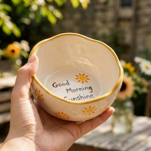Könnte beinhalten: Cremefarbene Keramikschale mit gewelltem Rand und goldfarbenem Rand. Die Schale hat ein handgemaltes Design aus orangefarbenen Sonnen und den Worten "Good Morning Sunshine" in schwarzer Schrift. Die Schale wird in einer Hand gehalten.