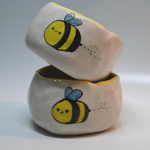 Könnte beinhalten: Zwei kleine, handgefertigte Keramikschalen, die übereinander gestapelt sind. Jede cremefarbene Schale hat einen gelben Rand und zeigt eine handgemalte Cartoon-Biene mit gelben und schwarzen Streifen und blauen Flügeln. Gestrichelte Linien deuten die Flugbahn der Biene an.