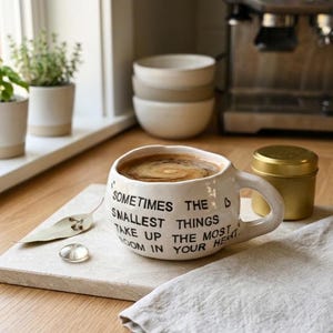 Könnte beinhalten: Eine weiße Keramik-Tasse gefüllt mit Kaffee, auf einem quadratischen, hellfarbigen Tablett. Die Tasse hat einen gebogenen Henkel und den Text "Sometimes the smallest things take up the most room in your heart." Ein kleiner goldener Behälter befindet sich in der Nähe.