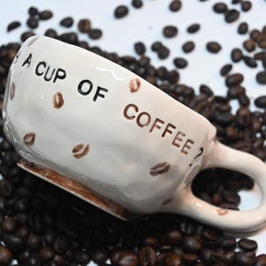 Könnte beinhalten: Cremefarbene Keramik-Kaffeetasse mit der Aufschrift "A CUP OF COFFEE?" und Kaffeebohnen-Illustrationen. Die Tasse ist von Kaffeebohnen umgeben, was eine warme, einladende Ästhetik erzeugt. Ein tolles Geschenk für Kaffeeliebhaber.