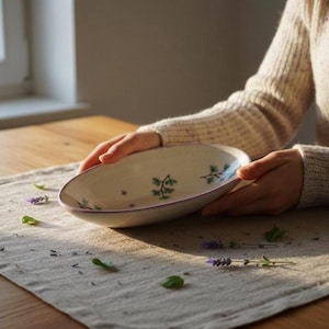 Könnte beinhalten: Ovale Servierschale aus Keramik mit cremefarbenem Grund und zartem lila Rand. Die Schale ist mit handgemalten grünen Blumenzweigen und kleinen lila Blumenakzenten verziert. Die Schale wird von einer Person auf einer Leinentischdecke gehalten.