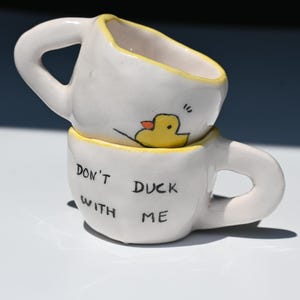 Könnte beinhalten: Zwei gestapelte weiße Keramiktassen mit unregelmäßigen, handgeformten Konturen. Die obere Tasse zeigt eine gelbe Entenillustration, während die untere Tasse den schwarzen Schriftzug "DON'T DUCK WITH ME" trägt. Beide Tassen haben gelbe Ränder.